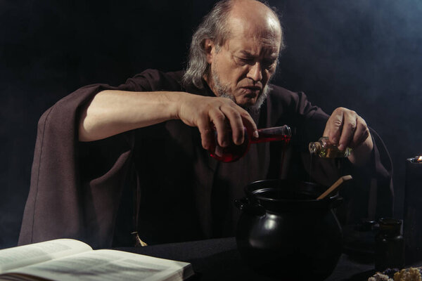 serious alchemist pouring liquid into pot near magic cookbook isolated on black