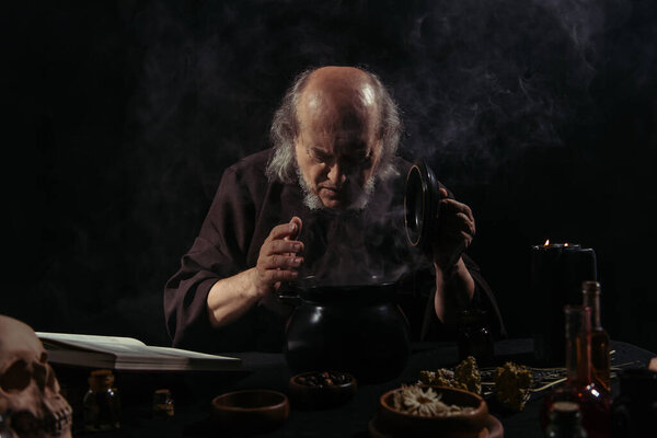 alchemist in dark robe looking into steaming pot near herbal ingredients on black background 