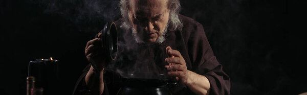 senior alchemist looking in boiling pot with potion isolated on black, banner
