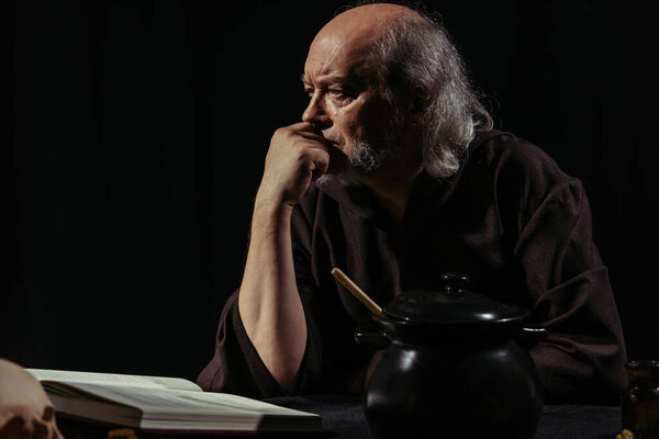 serious alchemist thinking near magic cookbook and pot isolated on black