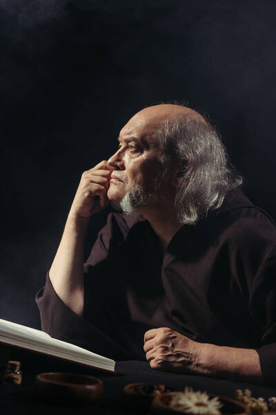 senior medieval alchemist thinking near magic cookbook isolated on black