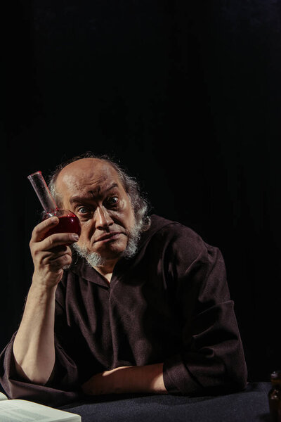 excited alchemist holding flask with magic potion and looking at camera isolated on black