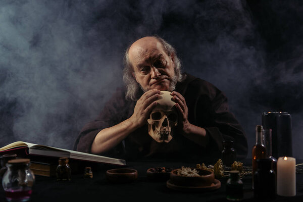 discouraged alchemist holding skull near ingredients and magic cookbook on black background with smoke