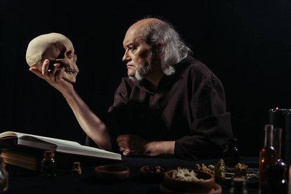 senior alchemist looking at skull near magic cookbook and herbal ingredients isolated on black