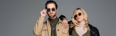 stylish man adjusting sunglasses near blonde woman in leather jacket isolated on grey, banner