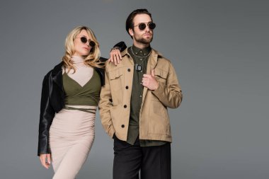 stylish man in sunglasses posing with hand in pocket near blonde woman in leather jacket and dress isolated on grey