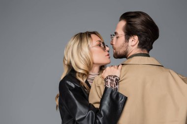 side view of blonde in stylish sunglasses hugging bearded man in beige jacket isolated on grey