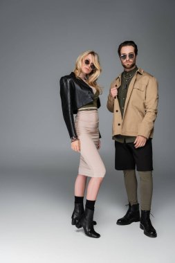 full length of stylish couple in autumnal outfits and sunglasses posing and looking at camera on grey