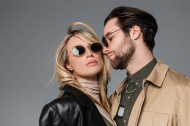 stylish bearded man in sunglasses looking at blonde woman in black leather jacket isolated on grey