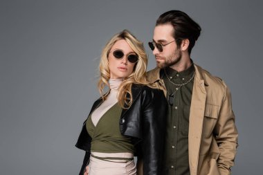 bearded man in sunglasses looking at blonde woman in black leather jacket isolated on grey