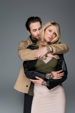 stylish bearded man in sunglasses embracing blonde woman in black leather jacket isolated on grey