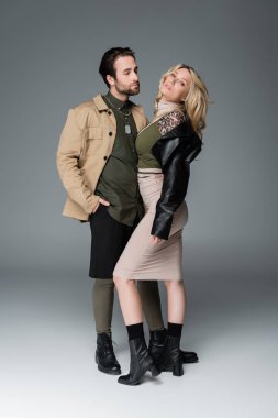 full length of stylish man in autumnal outfit posing with hand in pocket near blonde woman on grey
