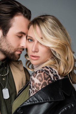stylish bearded man near blonde woman in trendy outfit looking at camera on grey