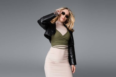 blonde woman in black leather jacket adjusting trendy sunglasses isolated on grey