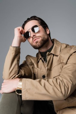bearded man in stylish jacket and sunglasses looking at camera isolated on grey