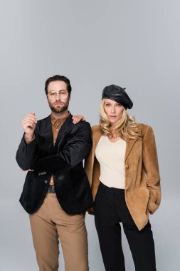 blonde woman in stylish beret posing with hand in pocket near bearded man in sunglasses isolated on grey