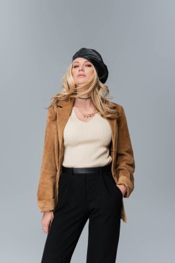 blonde woman in stylish leather beret and beige blazer standing with hand in pocket isolated on grey