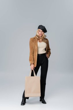 full length of blonde woman in stylish leather beret and beige blazer holding shopping bag on grey