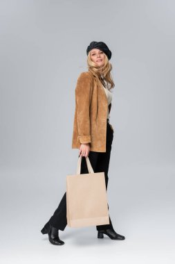 full length of blonde model in stylish leather beret and beige blazer holding shopping bag on grey