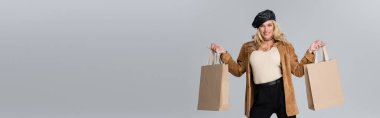 happy woman in stylish leather beret and beige blazer holding shopping bags isolated on grey, banner