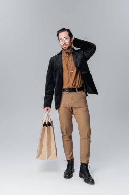 full length of bearded man in sunglasses and trendy autumnal outfit posing with shopping bags on grey