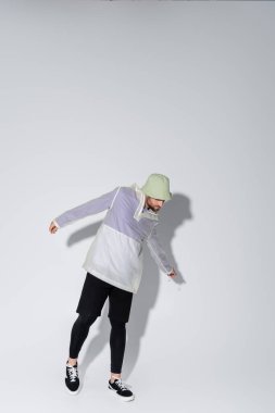 full length of young man in stylish sportive clothing and panama hat posing on grey