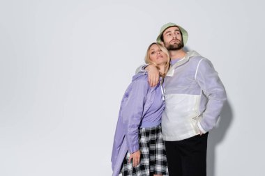 bearded man in panama hat standing with hand in pocket hugging blonde girlfriend in tartan skirt on grey