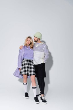 full length of bearded man in panama hat hugging stylish woman in tartan skirt and longs socks with sneakers on grey