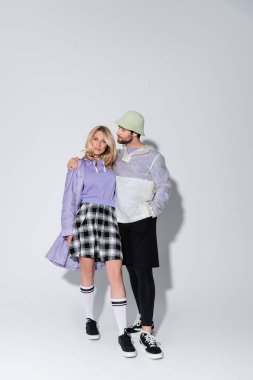 full length of bearded man in panama hat hugging stylish woman in tartan skirt and longs socks with sneakers on grey clipart