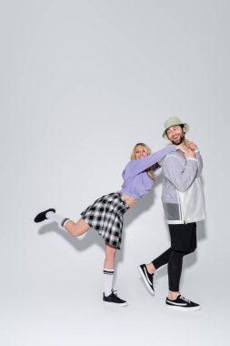 full length of happy woman in tartan skirt and longs socks with sneakers leaning on man in panama hat on grey clipart
