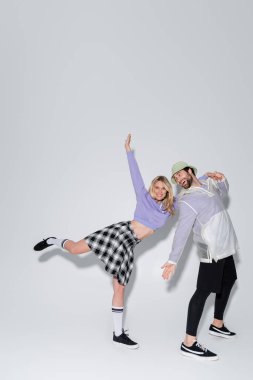 full length of cheerful woman in tartan skirt and longs socks with sneakers leaning on man in panama hat on grey