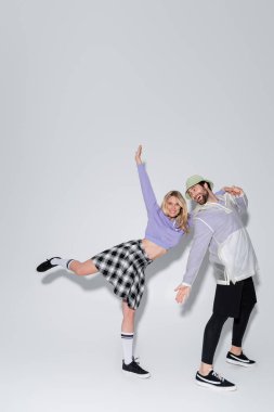 full length of cheerful woman in tartan skirt and longs socks with sneakers leaning on man in panama hat on grey clipart