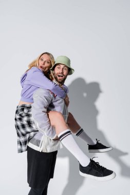 amazed man in panama hat piggybacking surprised woman in longs socks and sneakers on grey clipart