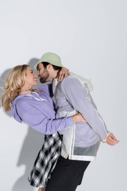 bearded man in panama hat kissing happy blonde girlfriend in tartan skirt on grey