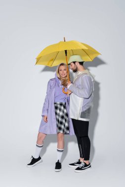 full length of woman in tartan skirt and longs socks standing with man in panama hat under yellow umbrella on grey clipart