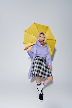 full length of blonde model in tartan skirt and longs socks with sneakers standing under yellow umbrella on grey