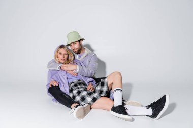 full length of stylish man in panama hat hugging blonde woman in tartan skirt and longs socks while sitting on grey