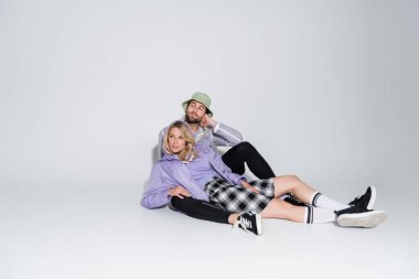 full length of man in stylish panama hat hugging blonde woman in tartan skirt and longs socks while sitting on grey