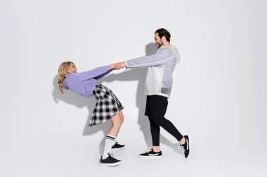 full length of happy man and blonde woman in tartan skirt holding hands on grey