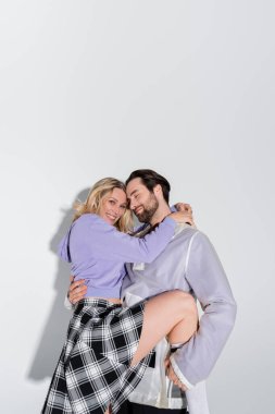 cheerful man holding in arms smiling blonde woman in tartan skirt on grey