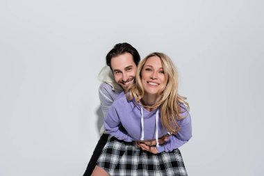 happy man hugging blonde woman in purple sweatshirt and tartan skirt while looking at camera on grey