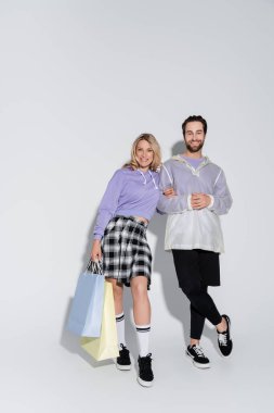 full length of happy and stylish couple holding shopping bags on grey