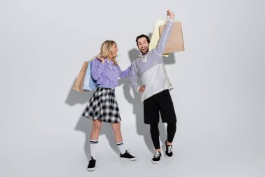 full length of excited man and cheerful woman in tartan skirt holding shopping bags on grey