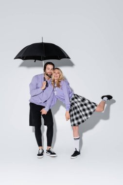 full length of happy couple in purple sweatshirts standing under umbrella on grey