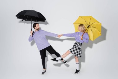 full length of happy couple in purple sweatshirts holding hands while standing under umbrellas on grey