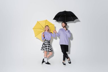 full length of pleased couple in purple sweatshirts holding hands while standing under umbrellas on grey clipart