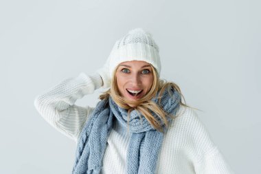 portrait of amazed woman in winter hat and knitted scarf looking at camera isolated on grey
