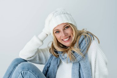 portrait of pleased woman in winter hat and knitted scarf looking at camera isolated on grey