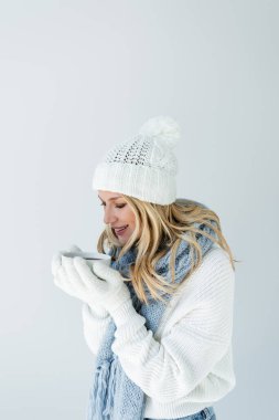 portrait of cheerful woman in winter hat and white gloves holding cup of hot beverage isolated on grey