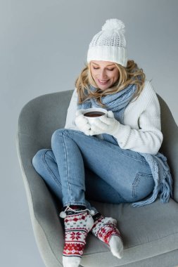 cheerful blonde woman in winter outfit sitting in comfortable armchair and holding cup of coffee isolated on grey